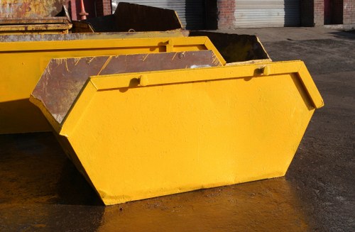 A large, rectangular yellow skip with reinforced steel edges and a smooth surface, positioned on a paved outdoor area. The skip is oriented horizontally, with its open top facing slightly to the right, revealing a spacious interior. The exterior shows signs of use, with some visible dirt and wear. Behind the yellow skip, parts of additional industrial containers or waste bins are partially visible, suggesting a commercial or industrial setting in Hounslow. The ground beneath the skip is dark and slightly damp, highlighting the outdoor environment. The image conveys a scene typical of waste collection or rubbish removal operations, with the skip ready for loading or transportation, and relates to the services provided by Commercial Waste Removal Hounslow in the local area.