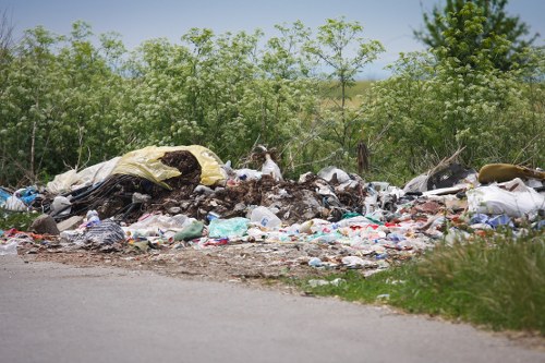 A large pile of mixed waste debris and rubbish, including plastic bags, clothing, and discarded materials, situated on the side of a road adjacent to a grassy area and small trees with green foliage. The waste appears uncontained and spread out over the pavement and verge, with some items partially covered by a yellow plastic sheet. The background features dense greenery and a clear sky, indicating an outdoor environment likely near a rural or suburban setting within Hounslow. The image reflects the type of unmanaged refuse that commercial waste removal services, such as those provided by Commercial Waste Removal Hounslow, are equipped to clear and dispose of professionally.