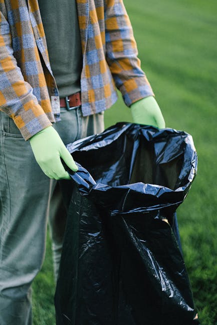 Three large black rubbish bags filled with waste material are placed on a paved sidewalk next to a black metal fence and some greenery, possibly in an urban or suburban setting. The bags appear to be 